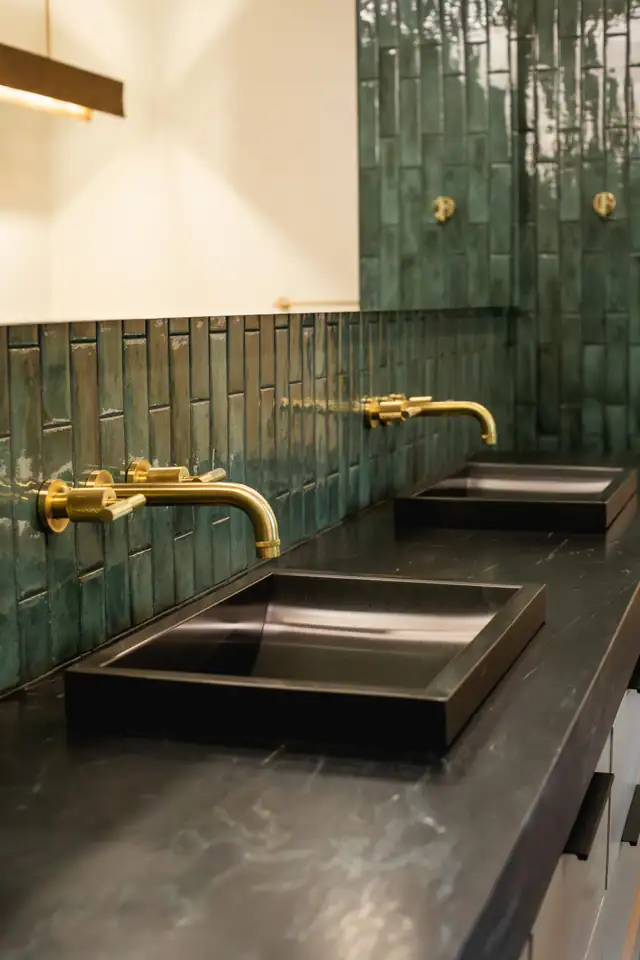 Black rectangular sinks with brass wall-mounted faucets against a green tile backsplash