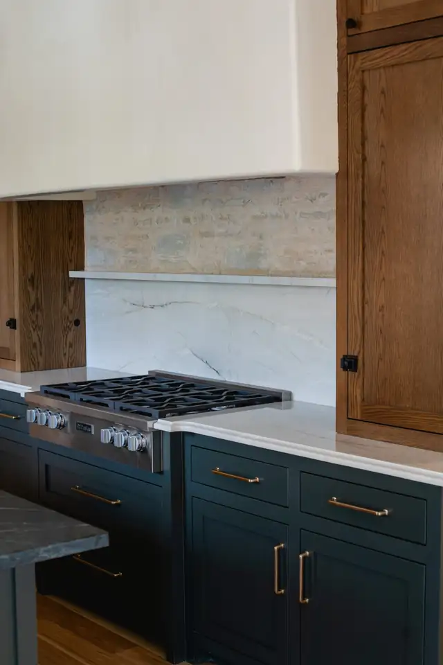 Kitchen with dark lower cabinets, white oak uppers, a stone slab backsplash and stainless gas range