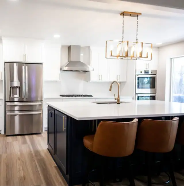 White kitchen by OKC Kitchen & Bath with a navy island, cognac leather counter stools, a brass chandelier and a stainless range hood