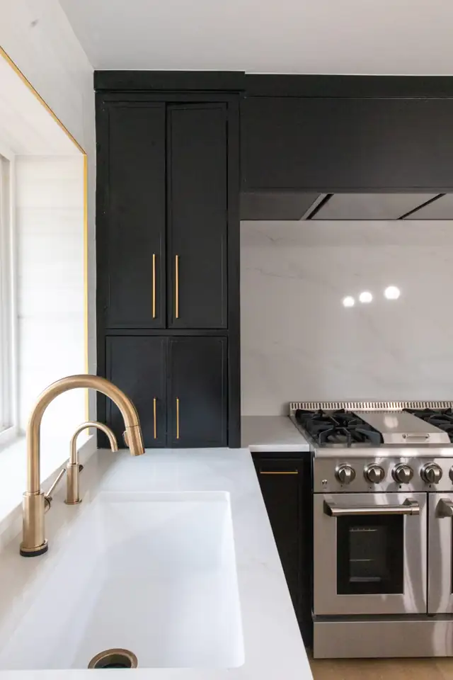 Black kitchen cabinets with brass pulls, a white quartz counter, a brass bridge faucet and a stainless range